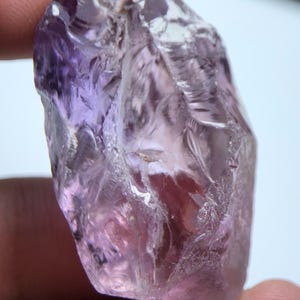 Könnte beinhalten: Ein roher Amethystkristall mit einer rauen, strukturierten Oberfläche. Der Kristall zeigt einen Farbverlauf von lilafarbenen Farbtönen, von tiefem Violett bis zu hellem Lavendel. Der Edelstein wird gehalten, was seine natürliche Form und Farbvariationen zeigt.