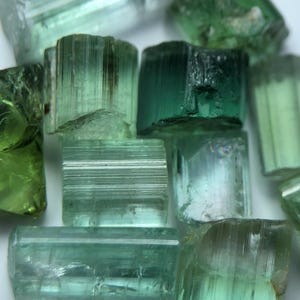 May include: Close-up of several raw, green tourmaline crystals. The crystals vary in shades from light to dark green, with visible striations and natural textures. The gemstones are irregularly shaped and appear translucent.