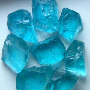 Könnte beinhalten: Acht rohe Aquamarin-Edelsteine in verschiedenen Größen und Formen. Die Kristalle sind von transluzenter hellblauer Farbe mit sichtbaren inneren Strukturen. Die raue, unregelmäßige Textur der Steine verleiht ihnen ein natürliches Aussehen.