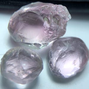 Könnte beinhalten: Drei rohe, ungeschliffene Edelsteine mit einem weichen, durchscheinenden rosa Farbton. Die Steine haben eine natürliche, unregelmäßige Form und eine leicht strukturierte Oberfläche, die das Licht reflektiert. Die Edelsteine scheinen eine ähnliche Größe zu haben und sind eng beieinander angeordnet.