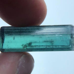 Könnte beinhalten: Ein rechteckiger, teal-farbener Turmalinkristall. Der Edelstein wird zwischen zwei Fingern gehalten und zeigt seine transluzente Qualität und natürliche Streifen. Die Farbe reicht von einem tiefen Teal bis zu einem helleren, fast transparenten Farbton mit sichtbaren inneren Einschlüssen.
