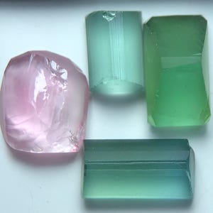 Pode incluir: Uma coleção de pedras preciosas brutas em tons de rosa, verde e azul-petróleo. As pedras preciosas têm várias formas, incluindo uma pedra rosa arredondada e vários cristais verdes e azul-petróleo retangulares e cilíndricos. As pedras são translúcidas.