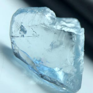 May include: A rough-cut, pale blue gemstone with a crystalline structure. The stone has a translucent quality, with visible internal fractures and a slightly irregular shape. The overall color is a soft, light blue, with areas of greater transparency.