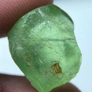Op de afbeelding: Een doorschijnende, lichtgroene edelsteen met een ruwe, onregelmatige vorm. De steen heeft een licht getextureerd oppervlak met donkere insluitsels en een klein, bruin, getextureerd gebied. De edelsteen wordt tussen twee vingers gehouden.
