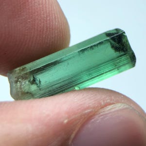 Könnte beinhalten: Ein durchscheinender, smaragdgrüner Turmalinkristall mit rechteckiger Form. Der Kristall hat eine leicht raue, strukturierte Oberfläche und einen dunklen, schwärzlichen Bereich an einem Ende. Der Kristall wird zwischen zwei Fingern vor weißem Hintergrund gehalten.