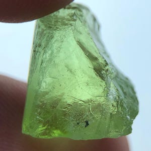 Könnte beinhalten: Ein durchscheinender, hellgrüner Peridot-Edelstein. Der grob geschliffene Stein hat eine kristalline Struktur und wird zwischen Fingern gehalten. Der Peridot hat ein natürliches, unpoliertes Aussehen mit sichtbaren Einschlüssen und einer leicht unebenen Oberfläche.