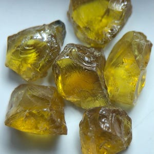 Könnte beinhalten: Eine Sammlung von sechs grob geschnittenen, durchscheinenden Edelsteinen. Die Steine haben einen goldgelben Farbton mit verschiedenen Bernstein- und Brauntönen. Die unregelmäßigen Formen und Facetten der Steine fangen das Licht ein.