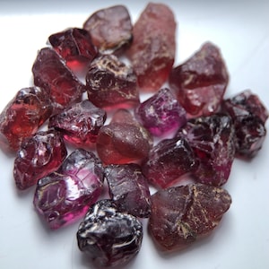 Könnte beinhalten: Eine Sammlung von rohen Granat-Edelsteinen in verschiedenen Rot- und Lilatönen. Die unpolierten Steine weisen natürliche Texturen und Facetten auf. Die Edelsteine sind auf einer weißen Oberfläche verstreut, was ihre natürliche Schönheit hervorhebt. Die Steine sind in verschiedenen Größen.