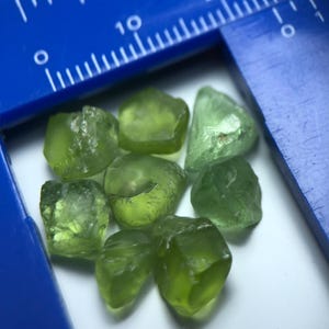 Könnte beinhalten: Acht rohe Peridot-Edelsteine in Olivgrün. Die Steine sind unregelmäßig geformt und durchscheinend. Ein blauer Lineal liegt im Hintergrund. Die Edelsteine liegen auf einer weißen Oberfläche.