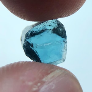 Könnte beinhalten: Ein kleiner, facettierter, blauer Edelstein, der zwischen zwei Fingern gehalten wird. Der Edelstein hat ein klares, kristallines Aussehen mit einem tiefblauen Farbton. Die Kanten sind rau, und das Licht reflektiert sich auf den Facetten.