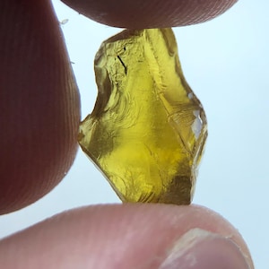 Puede incluir: Una gema translúcida de color amarillo dorado, de forma irregular, se sostiene entre dos dedos. La gema tiene una superficie rugosa y texturizada con patrones internos visibles y un ligero brillo. El fondo es blanco suave y desenfocado.