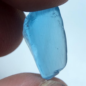Könnte beinhalten: Ein kleines, unregelmäßig geformtes Stück durchscheinendes, hellblaues Glas. Das Glas wird zwischen zwei Fingern vor einem weißen Hintergrund gehalten. Die Kanten des Glases sind rau und uneben.