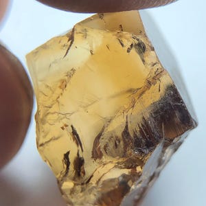 Könnte beinhalten: Ein grob geschliffener, durchscheinender Edelstein mit einem warmen, bernsteinfarbenen Farbton. Der Stein hat eine kristalline Struktur mit inneren Brüchen und dunklen Einschlüssen. Die Oberfläche reflektiert das Licht und zeigt seine natürliche Textur und Farbvariationen. Der Edelstein wird zwischen zwei Fingern gehalten.