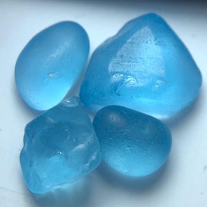 Peut inclure: Quatre morceaux de verre de mer de formes et tailles variées. Le verre est d'un bleu clair translucide, avec des bords lisses et arrondis. Les pièces sont disposées les unes près des autres, créant une collection de trésors naturels usés par l'océan.