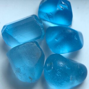 Könnte beinhalten: Fünf durchscheinende, hellblaue Edelsteine liegen auf einer weißen Oberfläche. Die Steine haben unterschiedliche Formen, einige sind facettierter, andere abgerundet. Die Farbe ist einheitlich, mit leichten Farbnuancen.