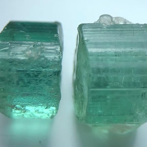 Könnte beinhalten: Zwei rohe, teal-farbene Turmalinkristalle. Die Kristalle haben eine rechteckige Form mit sichtbaren Streifen und einem durchscheinenden Aussehen. Die Kristalle sind vor einem weißen Hintergrund platziert, was ihre natürliche Schönheit hervorhebt.