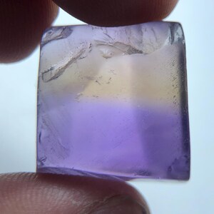 Könnte beinhalten: Ein quadratischer, durchscheinender Edelstein, der zwischen Fingern gehalten wird. Er zeigt einen Farbverlauf von blassgelb nach lavendel-violett. Die Oberfläche des Steins ist leicht strukturiert.