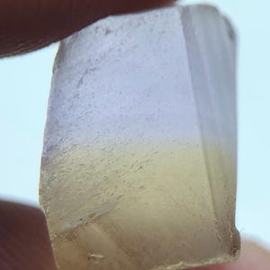Puede incluir: Un cristal rectangular translúcido con un degradado de colores. La parte superior es de un lavanda pálido, que pasa a un amarillo suave en la base. El cristal se sostiene entre los dedos, mostrando su forma natural y en bruto.