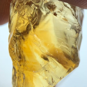 Könnte beinhalten: Nahaufnahme eines rauen, ungeschliffenen Edelsteins mit einem warmen, goldgelben Farbton. Der durchscheinende Stein hat eine kristalline Struktur mit sichtbaren inneren Brüchen und eine leicht raue, unebene Oberfläche. Der Edelstein wird von einem Finger gehalten.