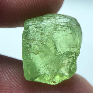 Könnte beinhalten: Ein roher, hellgrüner Peridot-Edelstein mit einer rauen, strukturierten Oberfläche. Der Edelstein wird zwischen zwei Fingern gehalten, was seine natürliche, unregelmäßige Form und seine durchscheinende Qualität zeigt. Der Peridot zeigt einen lebendigen, olivgrünen Farbton.