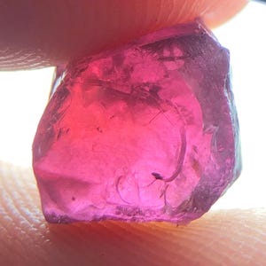 Könnte beinhalten: Ein transluzenter, facettierter, rosa Edelstein mit einer rauen, unregelmäßigen Form. Der Edelstein wird zwischen Fingern gehalten, wobei das Licht durchscheint und seine Farbe und innere Struktur hervorhebt. Die Farbe des Edelsteins reicht von tiefem Rosa bis zu einem helleren, transparenteren Farbton.