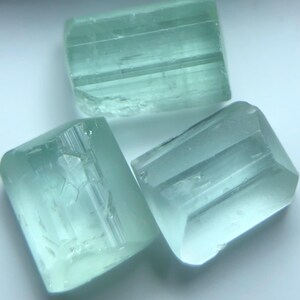 Puede incluir: Tres cristales de aguamarina en bruto, de color azul verdoso claro. Las piedras preciosas son rectangulares con estructuras internas y facetas visibles. Los cristales son translúcidos y están dispuestos sobre una superficie blanca.