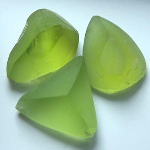 Könnte beinhalten: Drei durchscheinende, facettierte Edelsteine in hellgrünen Farbtönen. Die Formen umfassen eine abgerundete, unregelmäßige Form und eine Tropfenform sowie ein dreieckiges Stück. Die Edelsteine haben ein glattes, poliertes Aussehen.