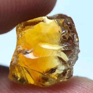 May include: A raw, golden-yellow gemstone held between two fingers. The gem is translucent, with internal fractures and an uneven surface. The colour varies from light amber to a deeper, honeyed tone. A natural, uncut mineral specimen.