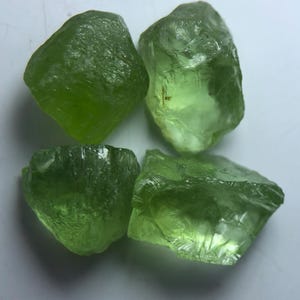Könnte beinhalten: Vier rohe, ungeschliffene Peridot-Edelsteine in verschiedenen Größen. Die Steine haben eine durchscheinende, leuchtend grüne Farbe mit rauen, strukturierten Oberflächen. Die Peridotkristalle werden vor einem weißen Hintergrund präsentiert.