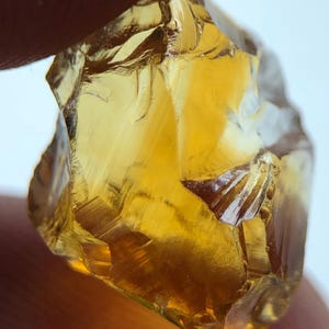 Könnte beinhalten: Nahaufnahme eines rauen, goldgelben Edelsteins. Der Edelstein hat eine kristalline Struktur mit mehreren Facetten, die das Licht reflektieren. Die Farbe ist ein sattes, warmes Gelb, wobei einige Bereiche durchscheinender erscheinen. Der Stein wird von einem Finger gehalten.