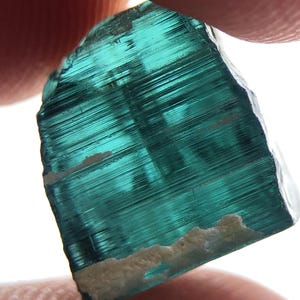 Puede incluir: Un cristal de turmalina translúcido de color verde azulado con estrías visibles. La gema tiene forma rectangular con una base rugosa de color beige. El cristal se sostiene entre dos dedos sobre un fondo blanco brillante.