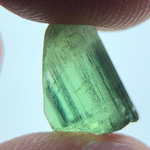 May include: A translucent, light green gemstone held between two fingers. The gem has a crystalline structure with visible striations and a slightly angled, triangular shape. The color is a vibrant, natural green.
