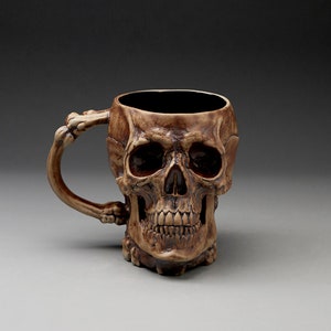 May include: A ceramic skull mug with a detailed design. The mug has a handle shaped like a bone and is a dark brown color.