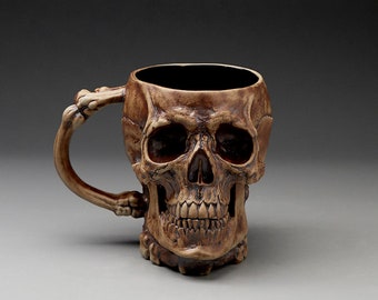 Puede incluir: Una taza de cerámica con forma de cráneo con un diseño detallado. La taza tiene un asa con forma de hueso y es de color marrón oscuro.