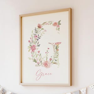 Puede incluir: Una impresión enmarcada con una ilustración de acuarela de la letra G rodeada de flores rosas y verdes. El nombre "Grace" está escrito en cursiva rosa debajo de la letra.