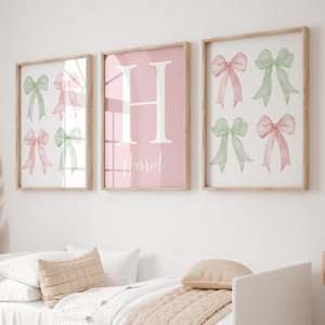 Puede incluir: Tres cuadros enmarcados. Dos presentan lazos de acuarela en rosa y verde. El marco central tiene un fondo rosa con la letra "H" y el nombre "Harriet" en escritura blanca. Los marcos son de madera clara.