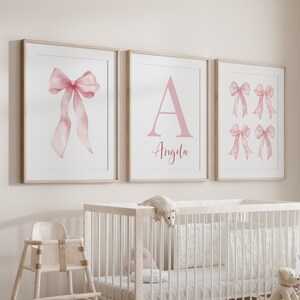 May include: Three framed prints with a nursery theme. The prints feature watercolor images of a pink bow, the letter "A" with the name "Angela", and four pink bows. The frames are light wood, and the background is a soft, neutral color.