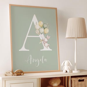 K&ouml;nnte beinhalten: Gerahmter Druck mit dem Buchstaben "A" und einem Hasen mit Ballons. Der Name "Angela" steht darunter. Der Hintergrund ist hellgr&uuml;n, der Rahmen hellbraun. Eine charmante Kinderzimmerdekoration.