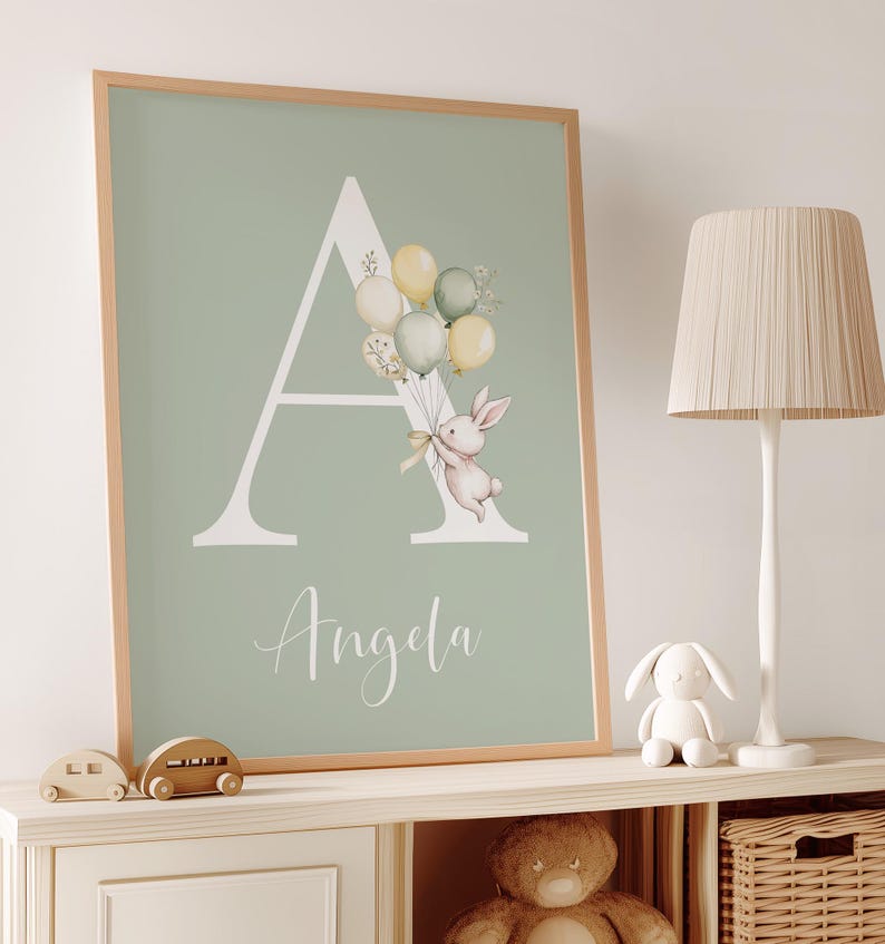 K&ouml;nnte beinhalten: Gerahmter Druck mit dem Buchstaben "A" und einem Hasen mit Ballons. Der Name "Angela" steht darunter. Der Hintergrund ist hellgr&uuml;n, der Rahmen hellbraun. Eine charmante Kinderzimmerdekoration.