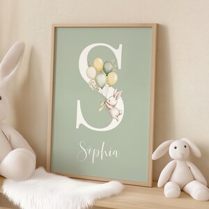 K&ouml;nnte beinhalten: Ein gerahmter Druck mit hellgr&uuml;nem Hintergrund, der einen gro&szlig;en wei&szlig;en Buchstaben "S" zeigt. Eine Aquarellillustration eines Hasen, der Ballons h&auml;lt, ist in den Buchstaben integriert. Der Name "Sophia" ist darunter in Kursivschrift geschrieben.
