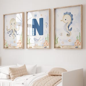 Puede incluir: Tres impresiones enmarcadas en acuarela con temática náutica. Una presenta un pulpo, otra la letra azul "N" con el nombre "Noah", y la tercera un caballito de mar. La obra incluye criaturas marinas, corales y algas, todo en colores pastel suaves.