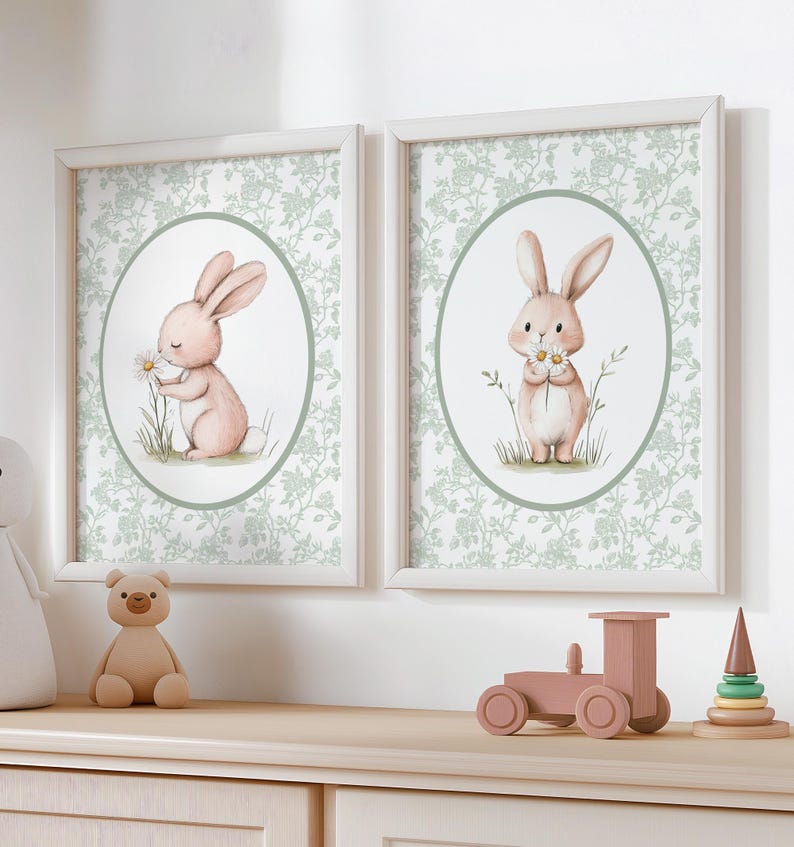 K&ouml;nnte beinhalten: Zwei gerahmte Drucke mit Aquarellillustrationen von rosa Hasen, die G&auml;nsebl&uuml;mchen halten. Die Hasen sind vor einem hellgr&uuml;nen floralen Hintergrund in einem ovalen Rahmen platziert. Die Drucke werden auf einer hellen Holzoberfl&auml;che zusammen mit einem Teddyb&auml;ren und Holzspielzeug ausgestellt.