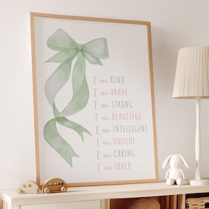 May include: A framed print featuring a watercolor illustration of a green bow with flowing ribbons. The print includes the text "I am KIND, BRAVE, STRONG, BEAUTIFUL, INTELLIGENT, UNIQUE, CARING, LOVED" in pink and green lettering.