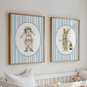 May include: Two framed prints with blue and white striped backgrounds. Each print features a circle with a cartoon bunny wearing clothes. One bunny is wearing a cap and the other is holding a lantern.