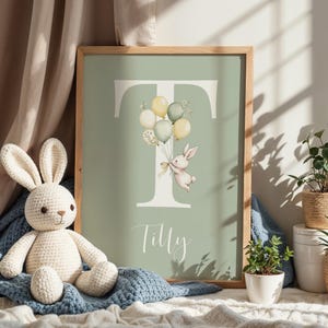 K&ouml;nnte beinhalten: Gerahmter Kunstdruck mit einem salbeigr&uuml;nen Hintergrund, der den Buchstaben "T" und eine Aquarellillustration eines Hasen mit Luftballons zeigt. Der Name "Tilly" steht darunter in Schreibschrift. Der Rahmen ist aus Holz.