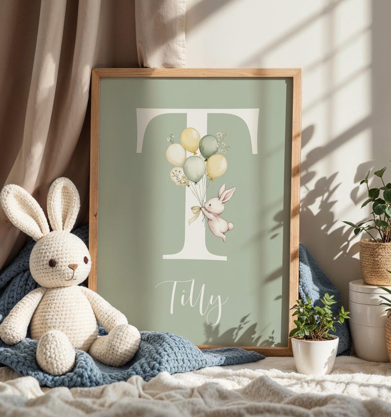 K&ouml;nnte beinhalten: Gerahmter Kunstdruck mit einem salbeigr&uuml;nen Hintergrund, der den Buchstaben "T" und eine Aquarellillustration eines Hasen mit Luftballons zeigt. Der Name "Tilly" steht darunter in Schreibschrift. Der Rahmen ist aus Holz.