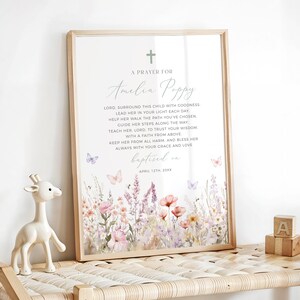 Op de afbeelding: Een ingelijste print met een bloemenontwerp en de tekst "A Prayer For Amelia Poppy". De print heeft een kruis, vlinders en een aquarel-achtige bloemenrand in tinten roze, paars en geel. Een houten giraffenbeeldje en houten blokken staan op de voorgrond.