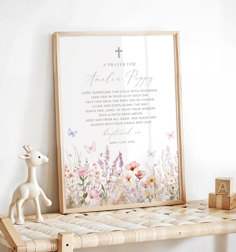 Op de afbeelding: Een ingelijste print met een bloemenontwerp en de tekst "A Prayer For Amelia Poppy". De print heeft een kruis, vlinders en een aquarel-achtige bloemenrand in tinten roze, paars en geel. Een houten giraffenbeeldje en houten blokken staan op de voorgrond.