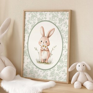 K&ouml;nnte beinhalten: Ein gerahmter Druck mit einer Aquarellillustration eines hellrosa Hasen, der G&auml;nsebl&uuml;mchen h&auml;lt. Der Hase befindet sich in einem ovalen Rahmen, vor einem floral gemusterten Hintergrund. Der Holzrahmen verleiht diesem charmanten Kunstwerk eine nat&uuml;rliche Note, ideal f&uuml;r die Kinderzimmerdekoration.