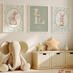 K&ouml;nnte beinhalten: Drei gerahmte Kunstdrucke f&uuml;r das Kinderzimmer mit Hasen und floralen Mustern. Ein Druck zeigt den Buchstaben "L" mit Luftballons und einem Hasen sowie den Namen "Lilly". Ein Pl&uuml;schelefant und sternf&ouml;rmige Kissen befinden sich in der N&auml;he eines Holzschranks.